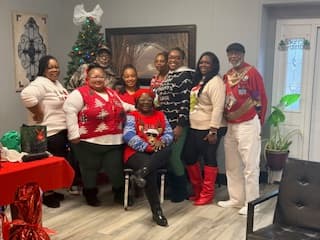 Group of people wearing festive Christmas sweaters and outfits pose together near a decorated tree.